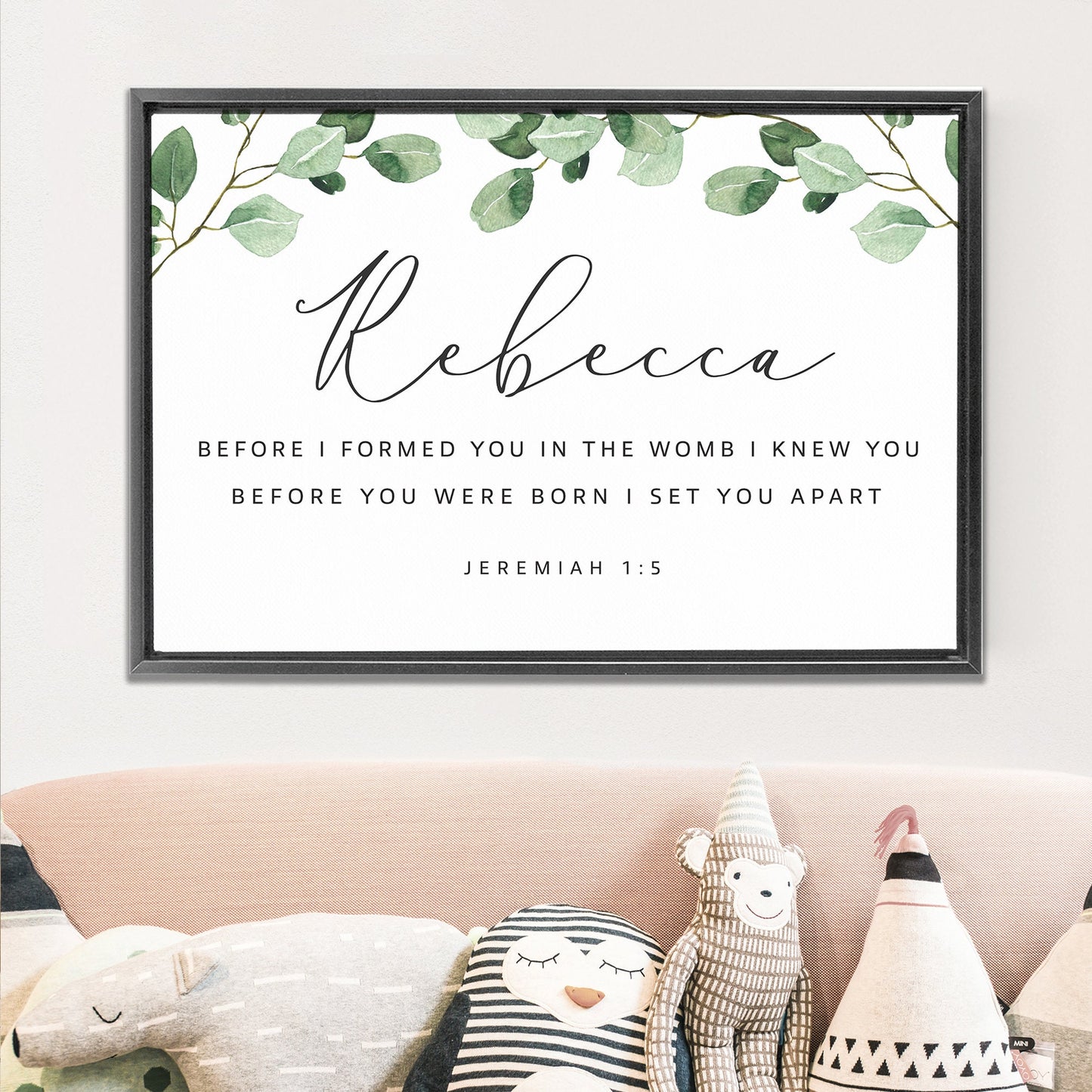 a framed Bible verse with the name Rebecca, along with a decorative border of green leaves. Below the Bible verse, there is a decorative border with a pattern of black and white stripes. The image also includes a stuffed animal and a pillow, adding a cozy and homey touch to the scene.