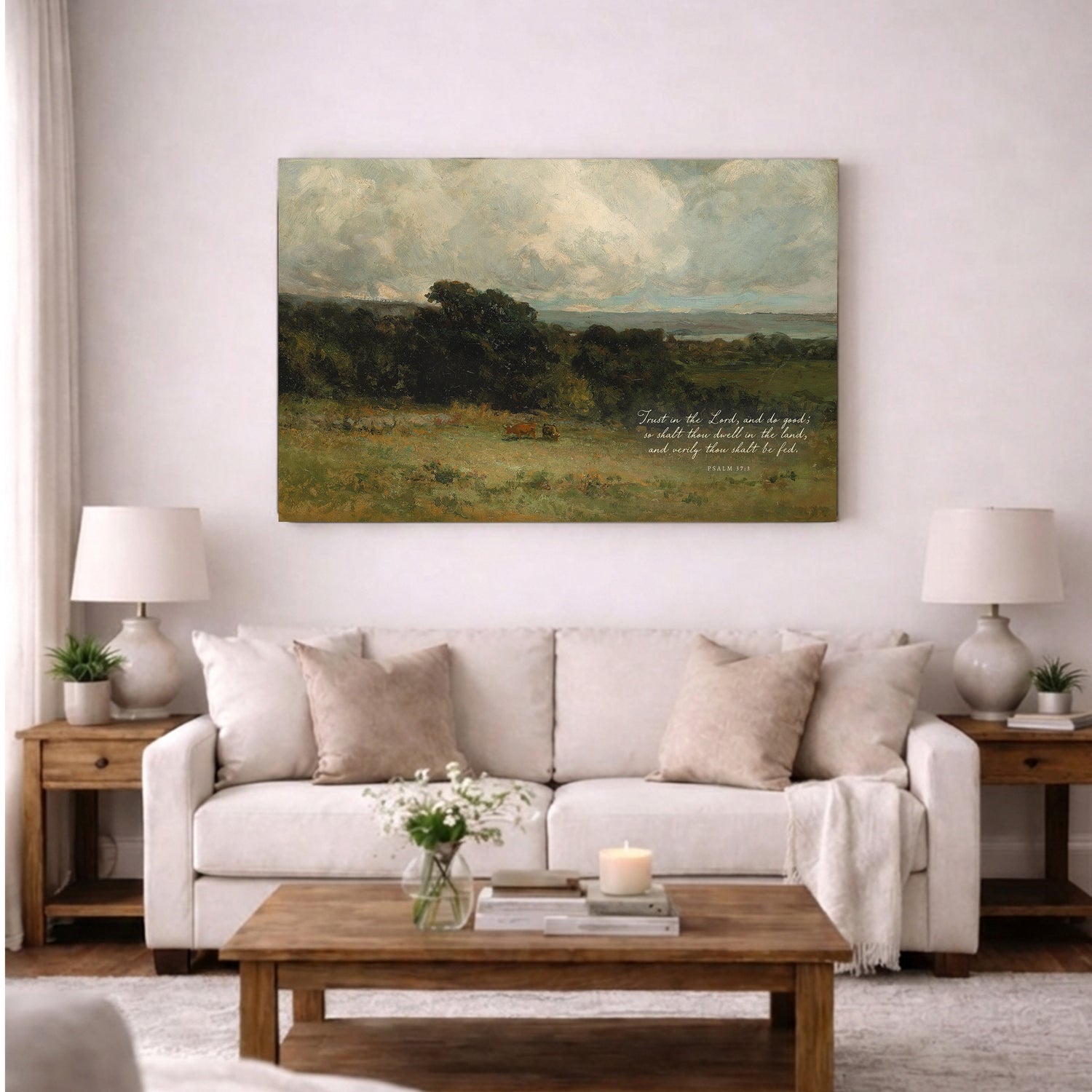 a living room with a large painting of a countryside landscape hanging on the wall above a white sofa. The sofa is adorned with throw pillows and a vase of flowers. In front of the sofa, there is a wooden coffee table with a book and a candle on it. The room also includes two lamps on either side of the sofa, and a window with curtains.