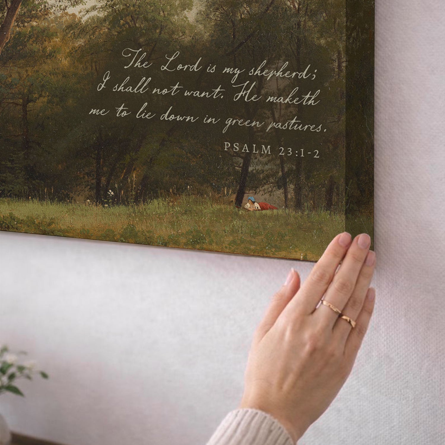 A hand is holding a framed print of a pastoral scene, with a Bible verse from Psalm 23:1-2 visible on the back.