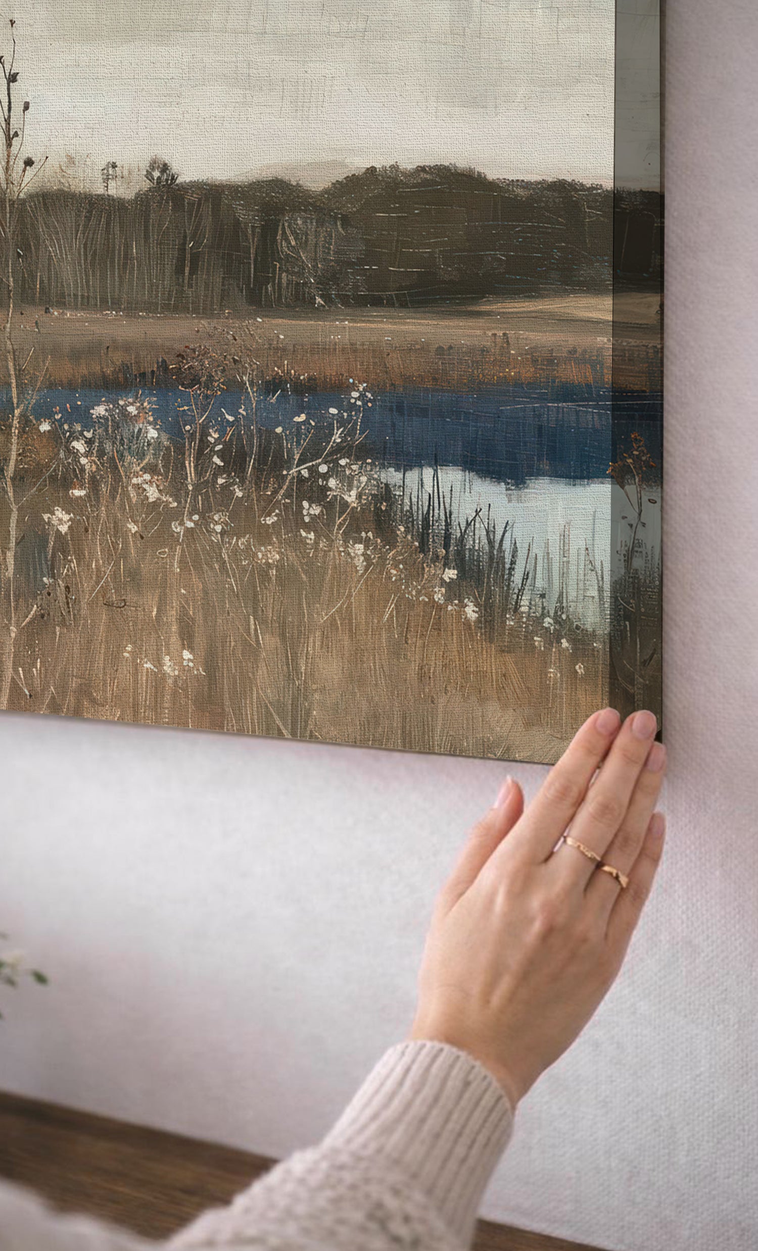 A person's hand is holding up a framed painting of a serene landscape, featuring a river, grassy field, and trees.