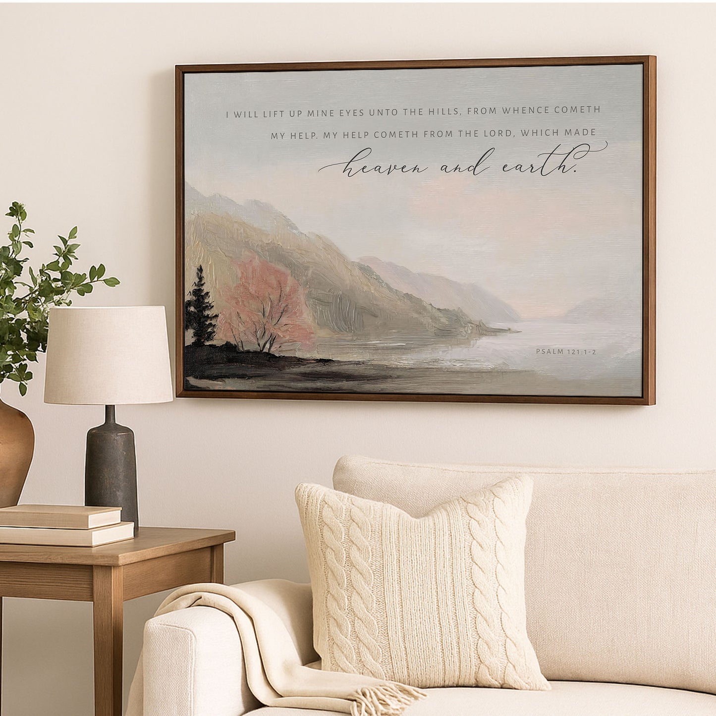 a framed painting of a serene landscape with mountains, trees, and a lake, hanging on a wall above a couch and a side table with a lamp.