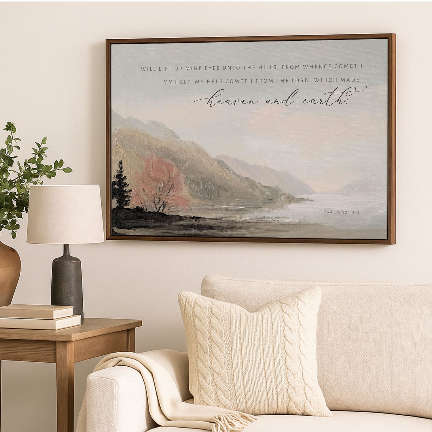 a framed painting of a serene landscape with mountains, trees, and a lake, hanging on a wall above a couch and a side table with a lamp.