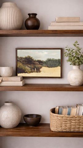 a wooden bookshelf with various decorative items and books arranged on the shelves. The bookshelf has three shelves, and there are several vases, bowls, and a plant displayed on the shelves. A framed landscape painting is hanging on the wall above the bookshelf, and a basket of books is placed on the lower shelf.