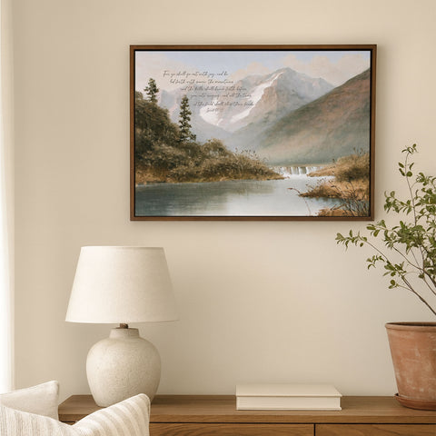 a serene landscape with a calm lake, surrounded by mountains and lush greenery. The painting is hung on a wall above a wooden table, which has a lamp and a plant on it.