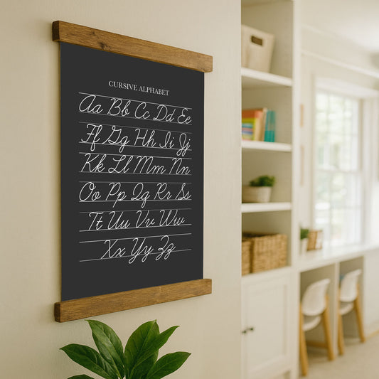 a blackboard with a cursive alphabet poster hanging on a wall, and there is a plant in the foreground.