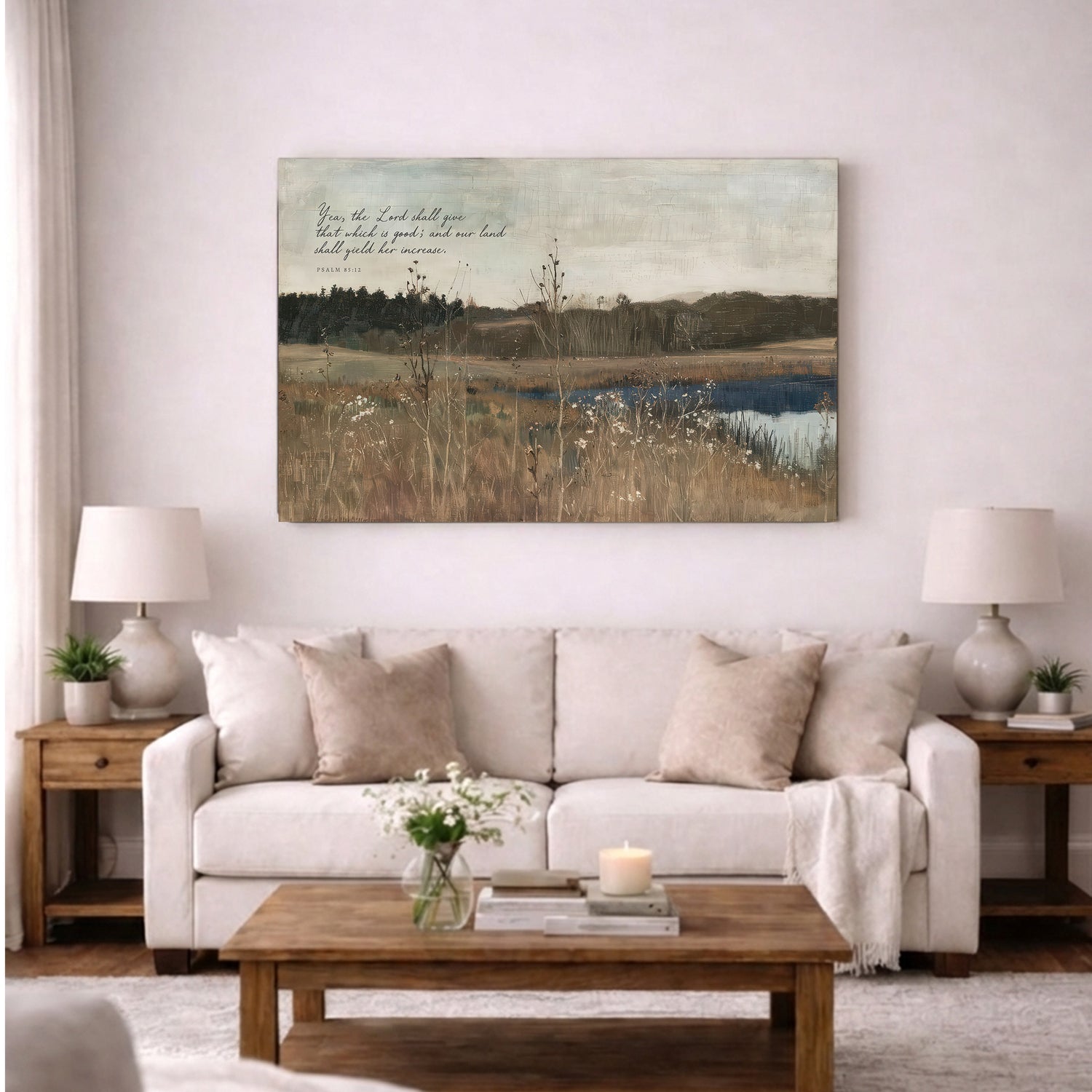 a living room with a large painting of a field and a body of water hanging on the wall above a white sofa. The sofa is adorned with pillows and a blanket, and there are two lamps on either side of the sofa. A wooden coffee table is placed in front of the sofa, and a vase of flowers is also present on the table.