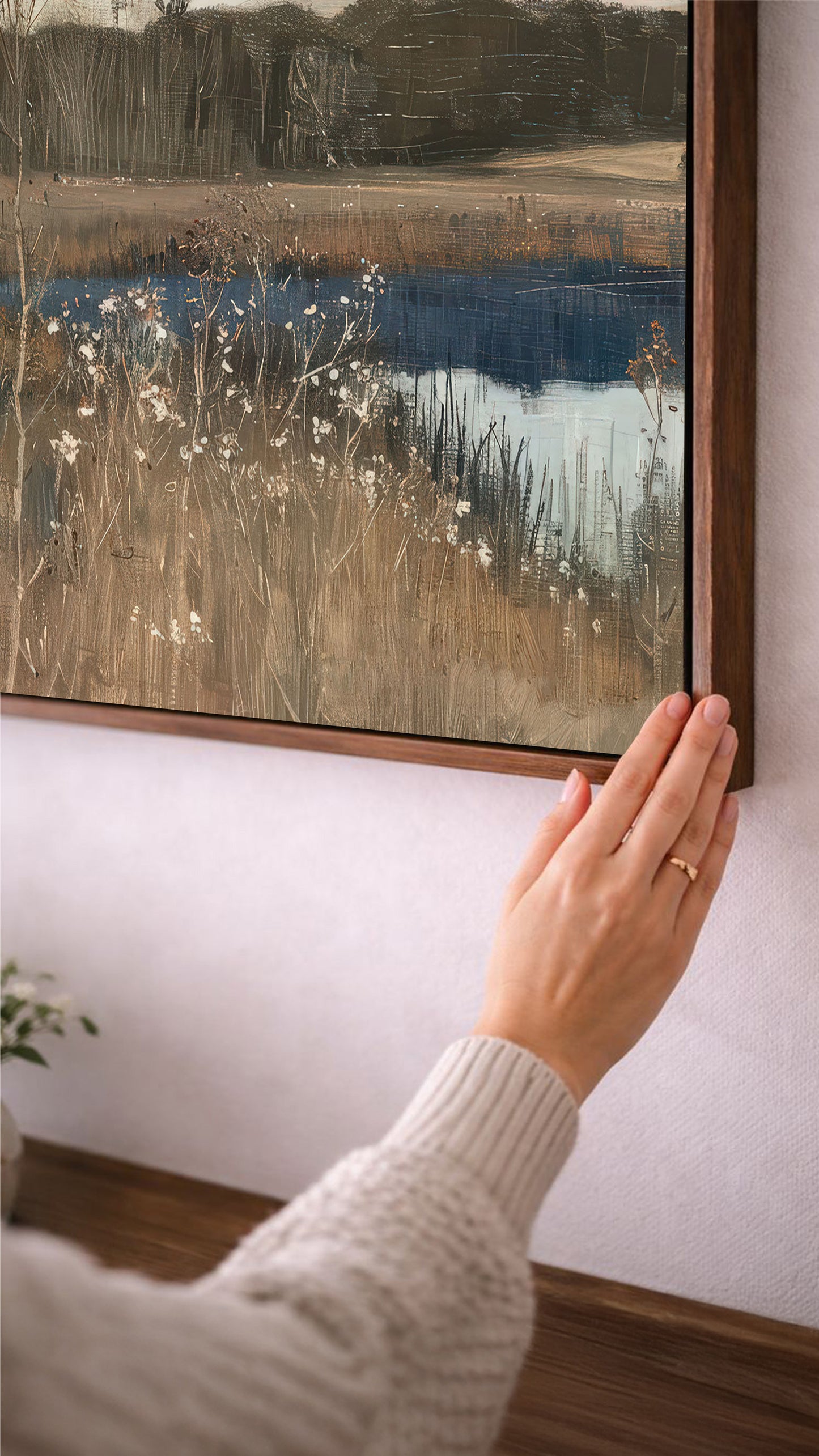 A person's hand is reaching towards a framed landscape painting on a wall, which depicts a serene countryside scene with a body of water and tall grass.
