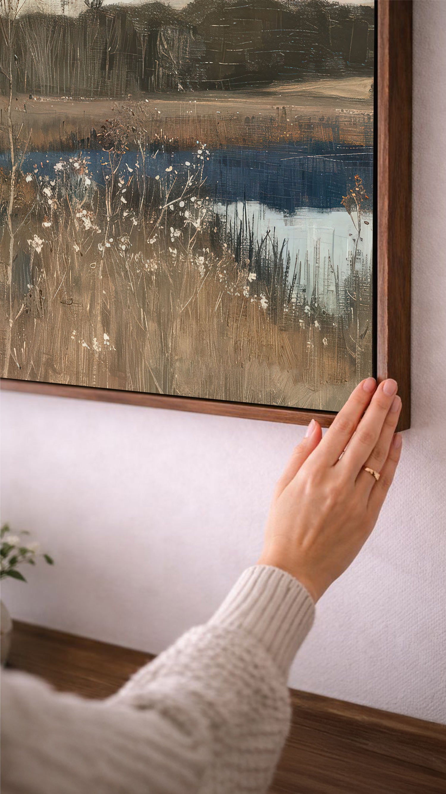 A person's hand is reaching towards a framed landscape painting on a wall, which depicts a serene countryside scene with a body of water and tall grass.