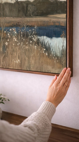 A person's hand is reaching towards a framed landscape painting on a wall, which depicts a serene countryside scene with a body of water and tall grass.