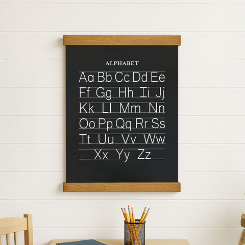 a blackboard with the alphabet written on it, hanging on a white wall. Below the blackboard, there is a metal pencil holder with pencils inside.