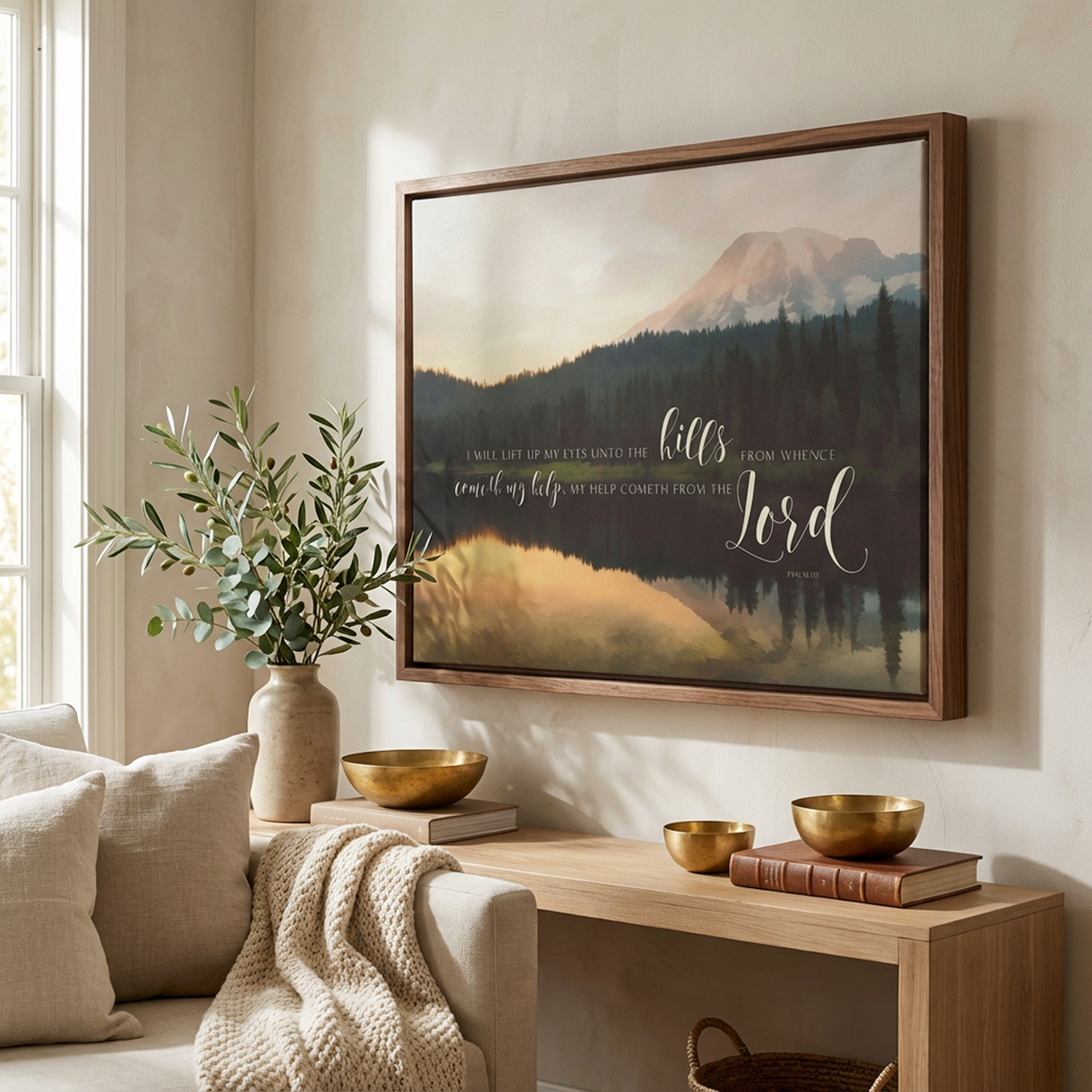 a living room with a large framed painting of a mountain landscape hanging on the wall. The painting has a serene and peaceful atmosphere, with the mountains and trees depicted in a realistic style. The room is decorated with a beige sofa, a wooden side table, and a vase with greenery. There are also two bowls and a book on the side table, adding to the cozy and inviting ambiance of the space.