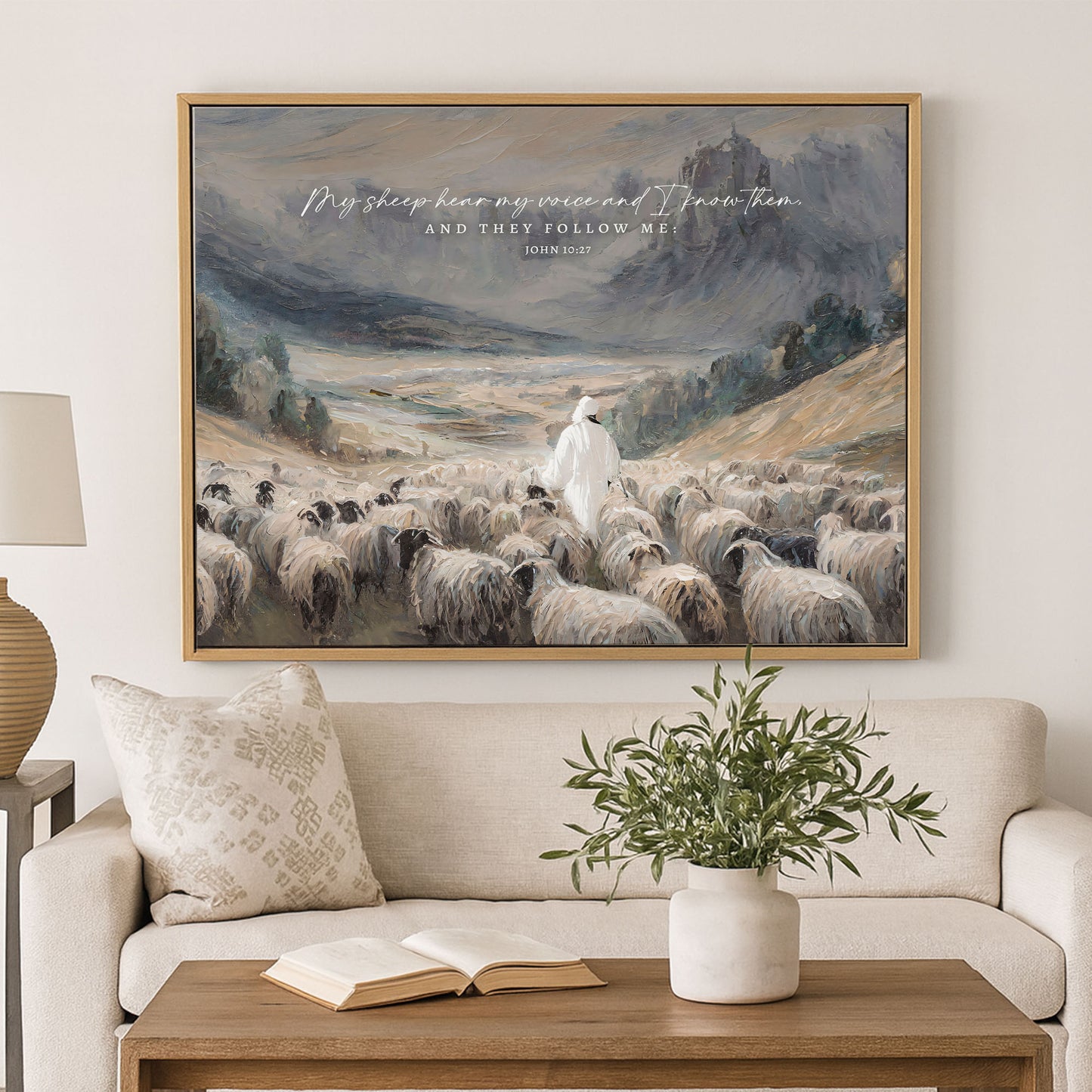 a large painting of a shepherd tending to his flock of sheep, with a mountainous landscape in the background. The painting is hung on a wall above a couch, which has a book and a vase on it.