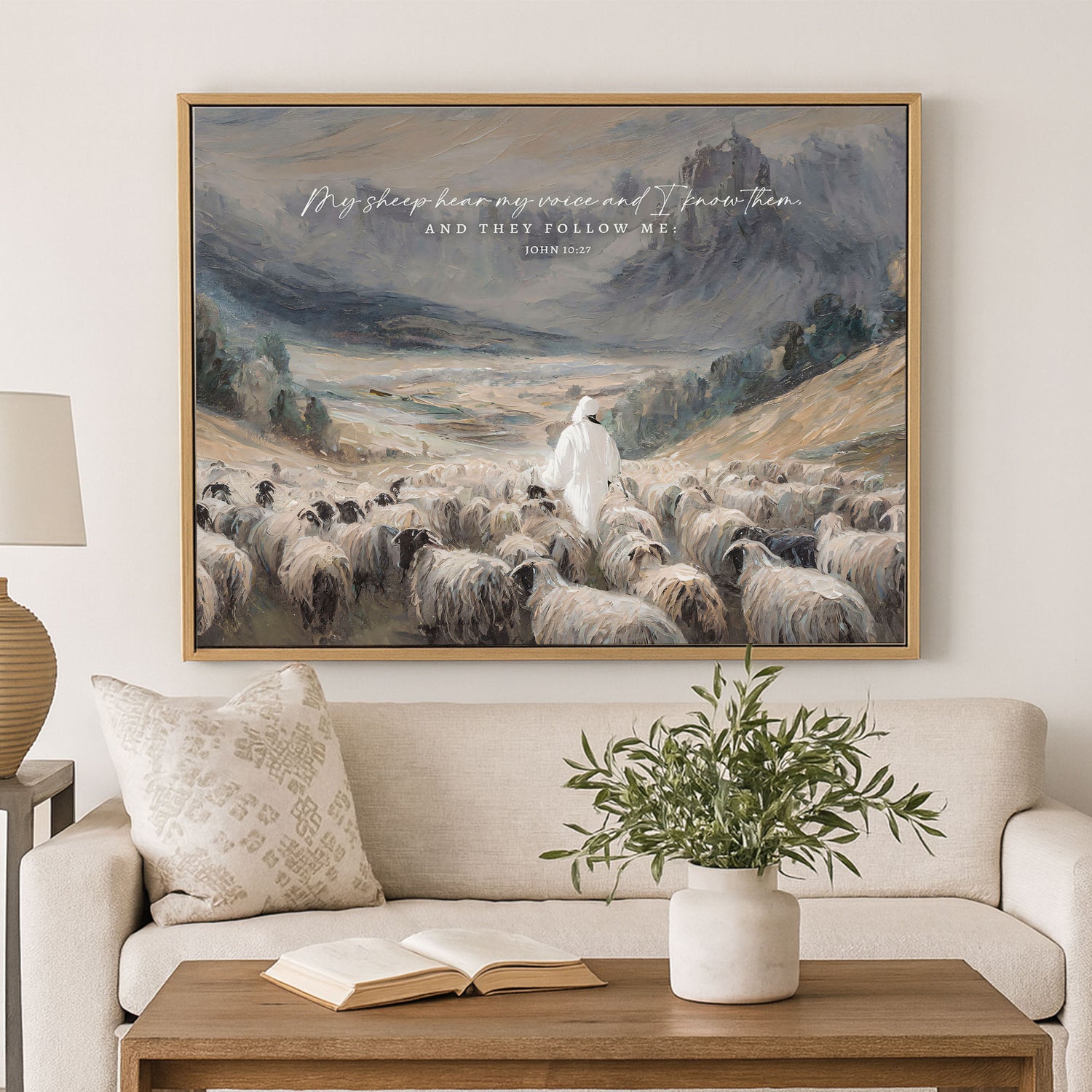 a large painting of a shepherd tending to his flock of sheep, with a mountainous landscape in the background. The painting is hung on a wall above a couch, which has a book and a vase on it.