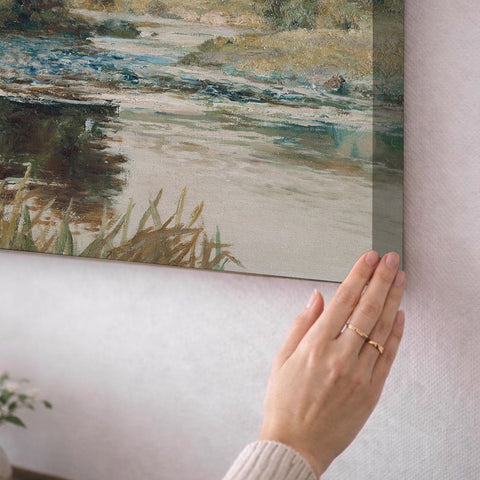 A person's hand is holding up a framed painting of a landscape, which features a river and grassy area.
