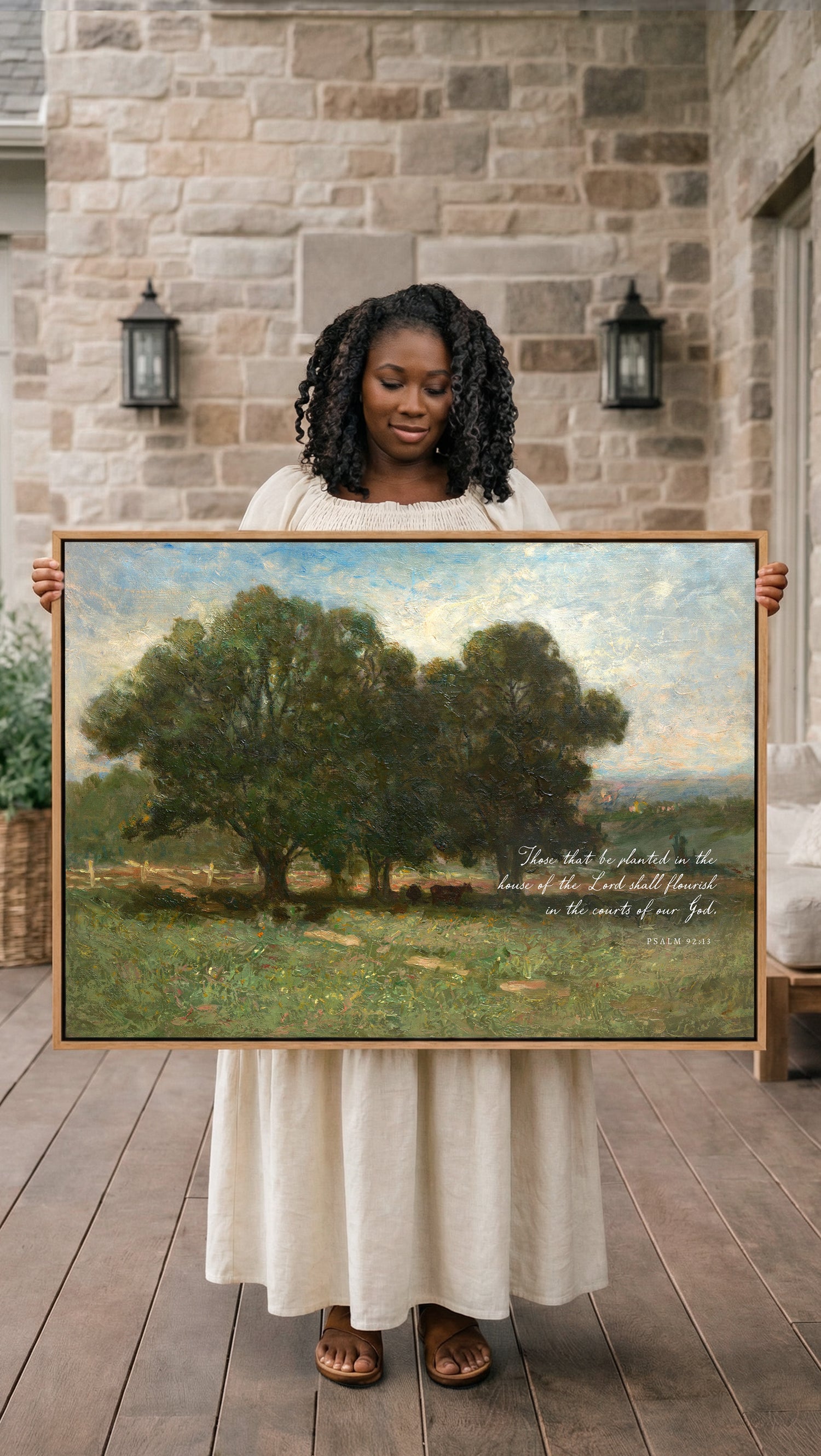 A woman in a white dress holds a large painting of a serene landscape with trees and a field, standing in front of a stone wall with lanterns.