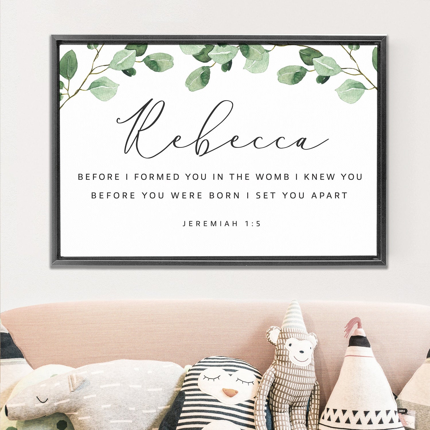 a framed Bible verse with the name Rebecca, along with a decorative border of green leaves. Below the Bible verse, there is a decorative border with a pattern of black and white stripes. The image also includes a stuffed animal and a pillow, adding a cozy and homey touch to the scene.