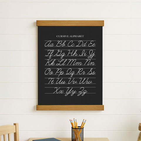 a blackboard with the cursive alphabet written on it, hanging on a white wall. Below the blackboard, there is a metal container filled with pencils.