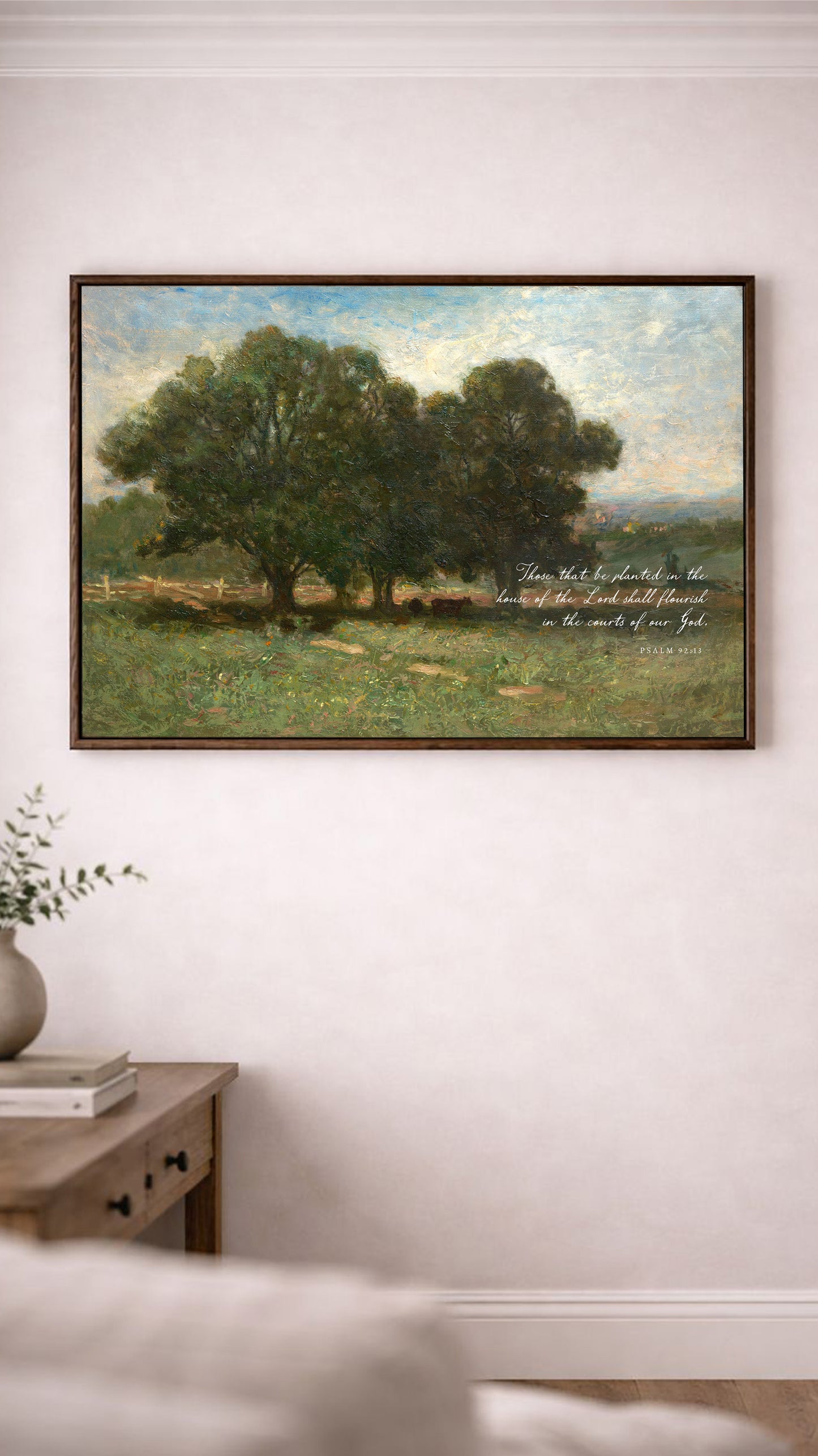 a painting of a serene landscape with a group of trees in the foreground and a peaceful countryside in the background. The painting is hung on a white wall above a wooden table with a vase of flowers on it.