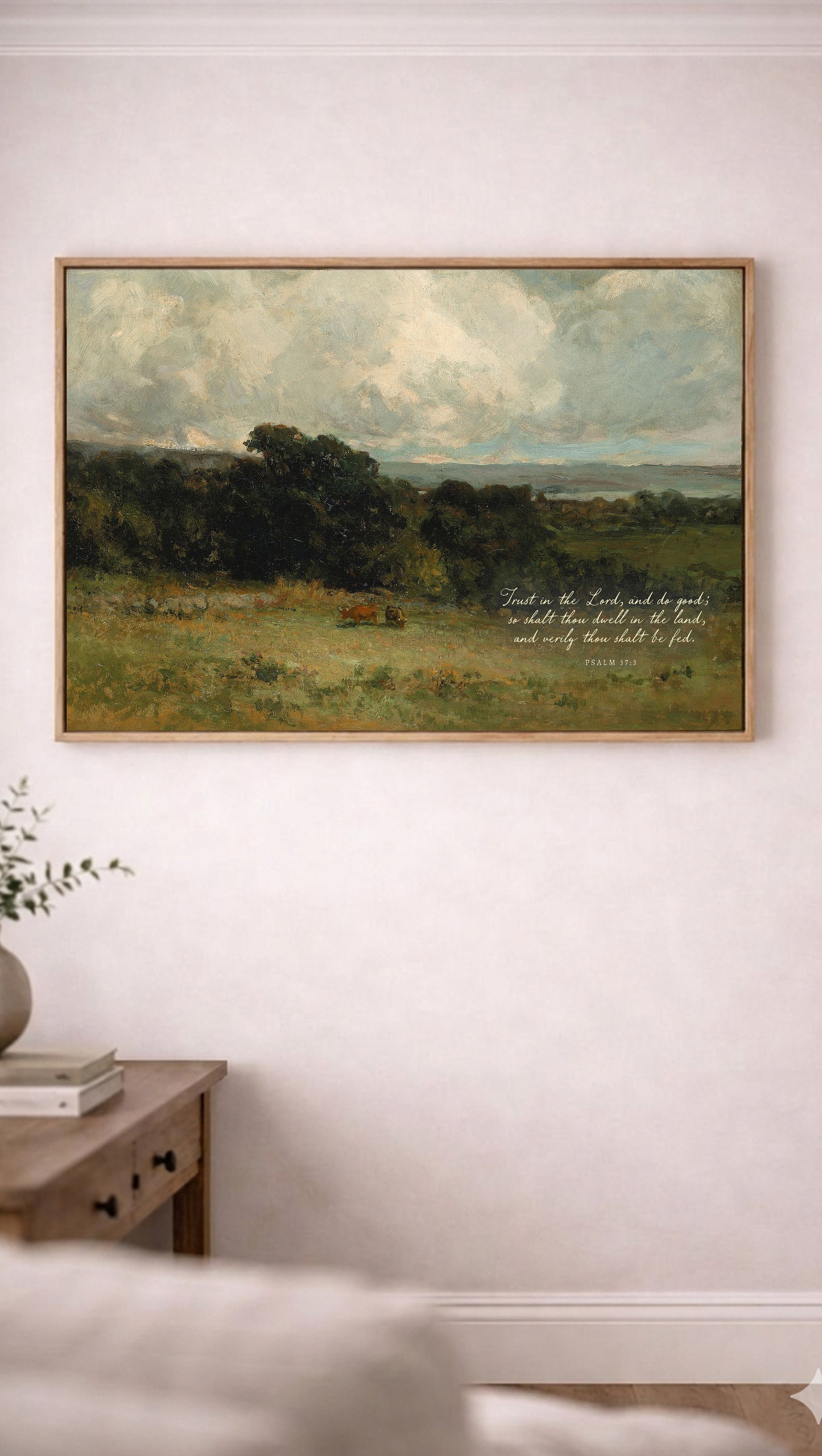 a painting of a pastoral landscape with a cow grazing in the foreground, hanging on a white wall above a wooden table with a plant on it.