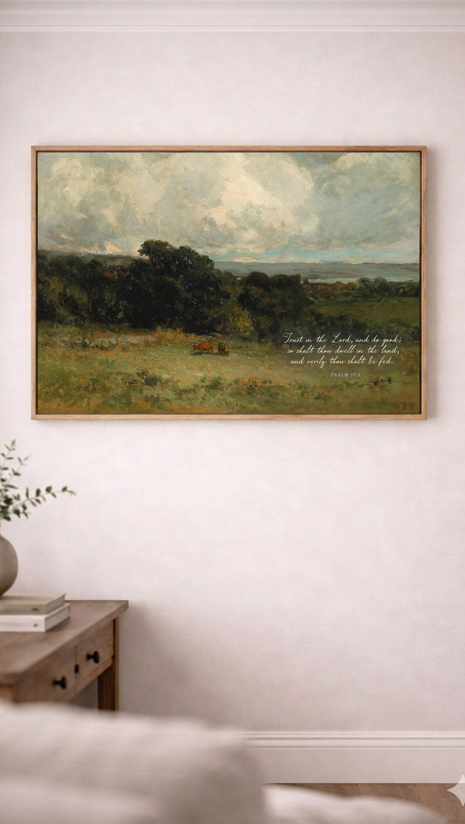 a painting of a pastoral landscape with a cow grazing in the foreground, hanging on a white wall above a wooden table with a plant on it.