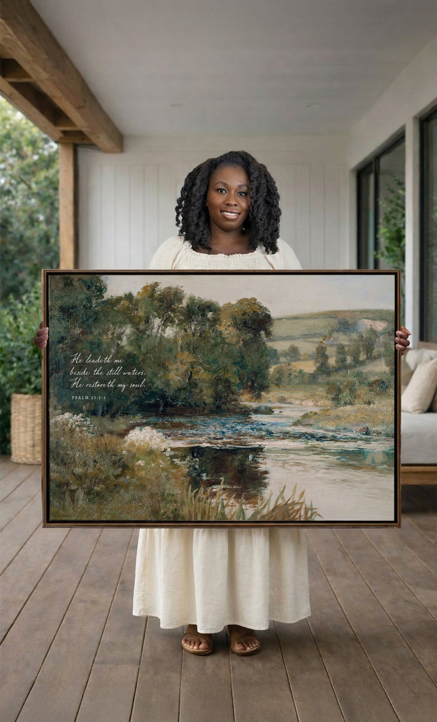 A woman in a white dress holds a large painting of a serene landscape with a river and trees, standing on a porch with a wooden floor.