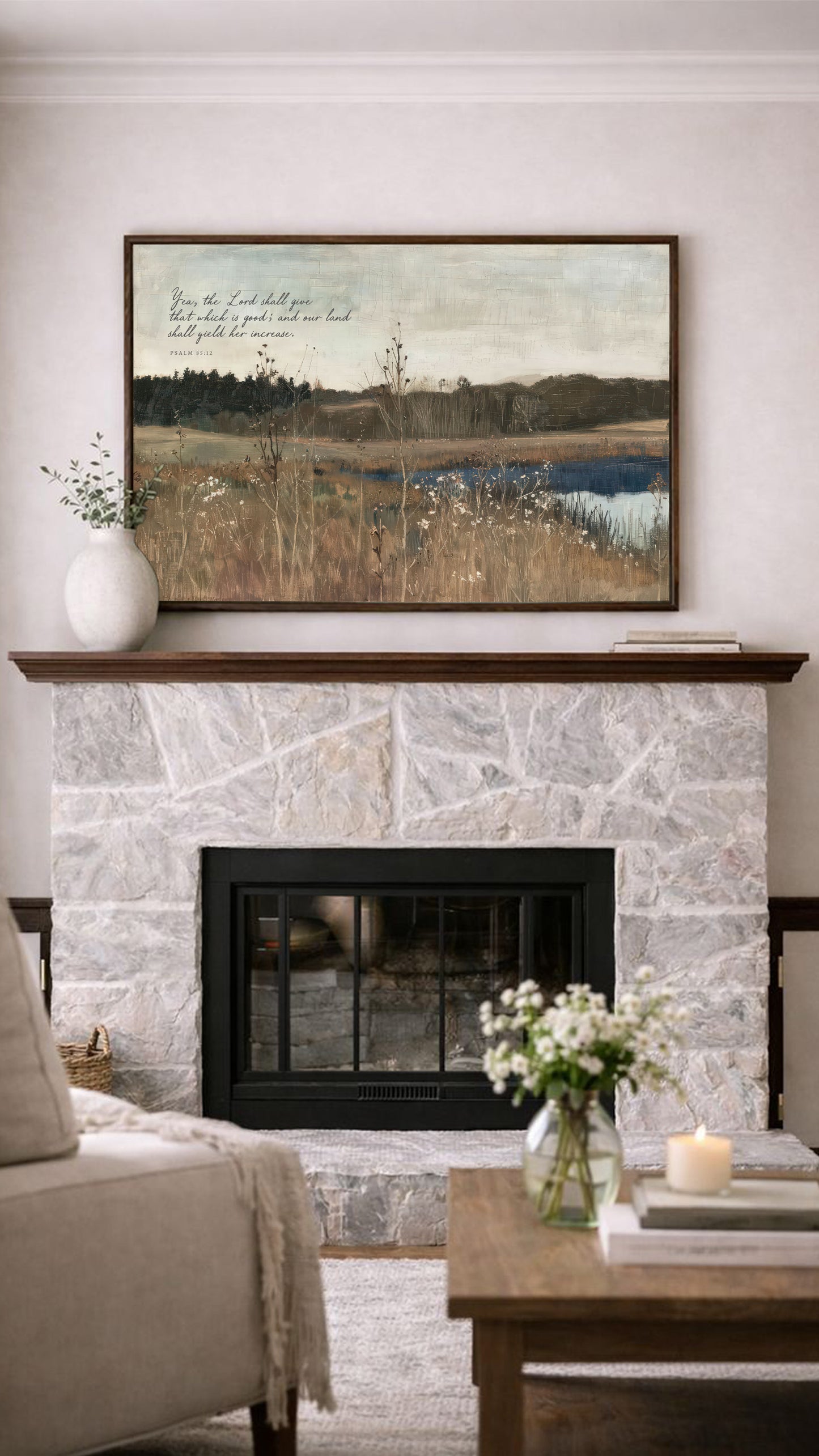 a cozy living room with a stone fireplace, a wooden coffee table, and a framed landscape painting above the fireplace. The painting depicts a serene countryside scene with tall grass and a body of water. The room is decorated with a vase of flowers, a candle, and a book, creating a warm and inviting atmosphere.