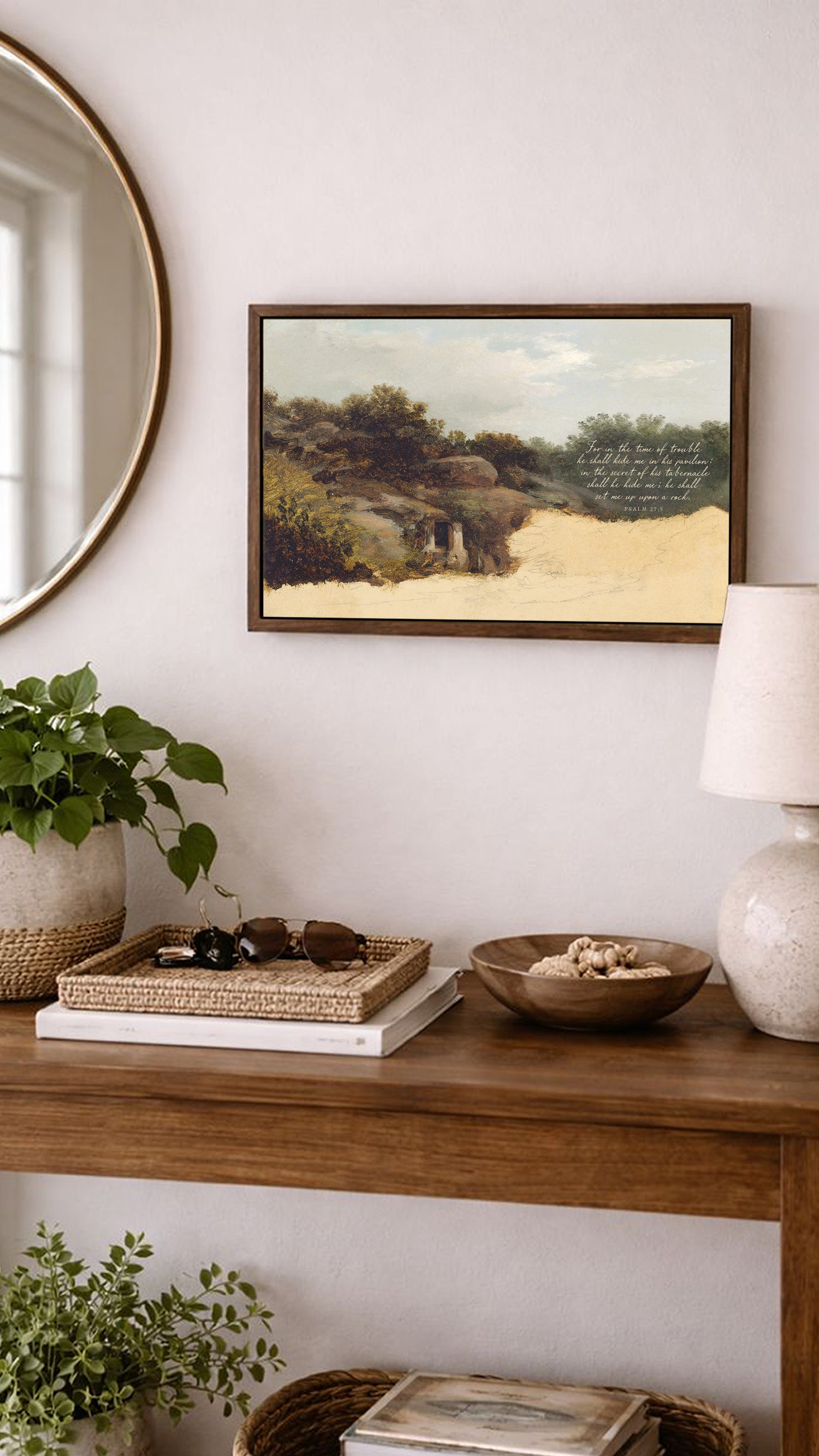 a wooden console table with various items on it, including a framed landscape painting, a lamp, a bowl, and a plant. The table is set against a white wall, and there is a round mirror above it.