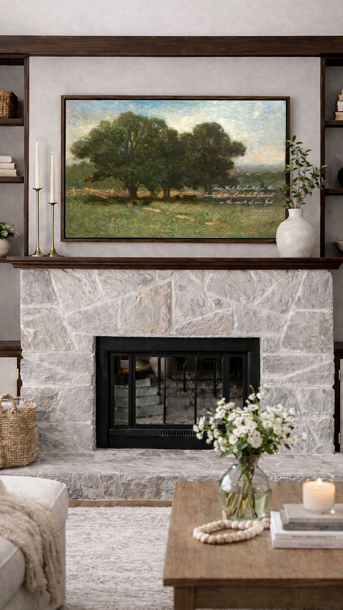 a cozy living room with a stone fireplace, a framed landscape painting above it, and a wooden coffee table with a vase of white flowers.
