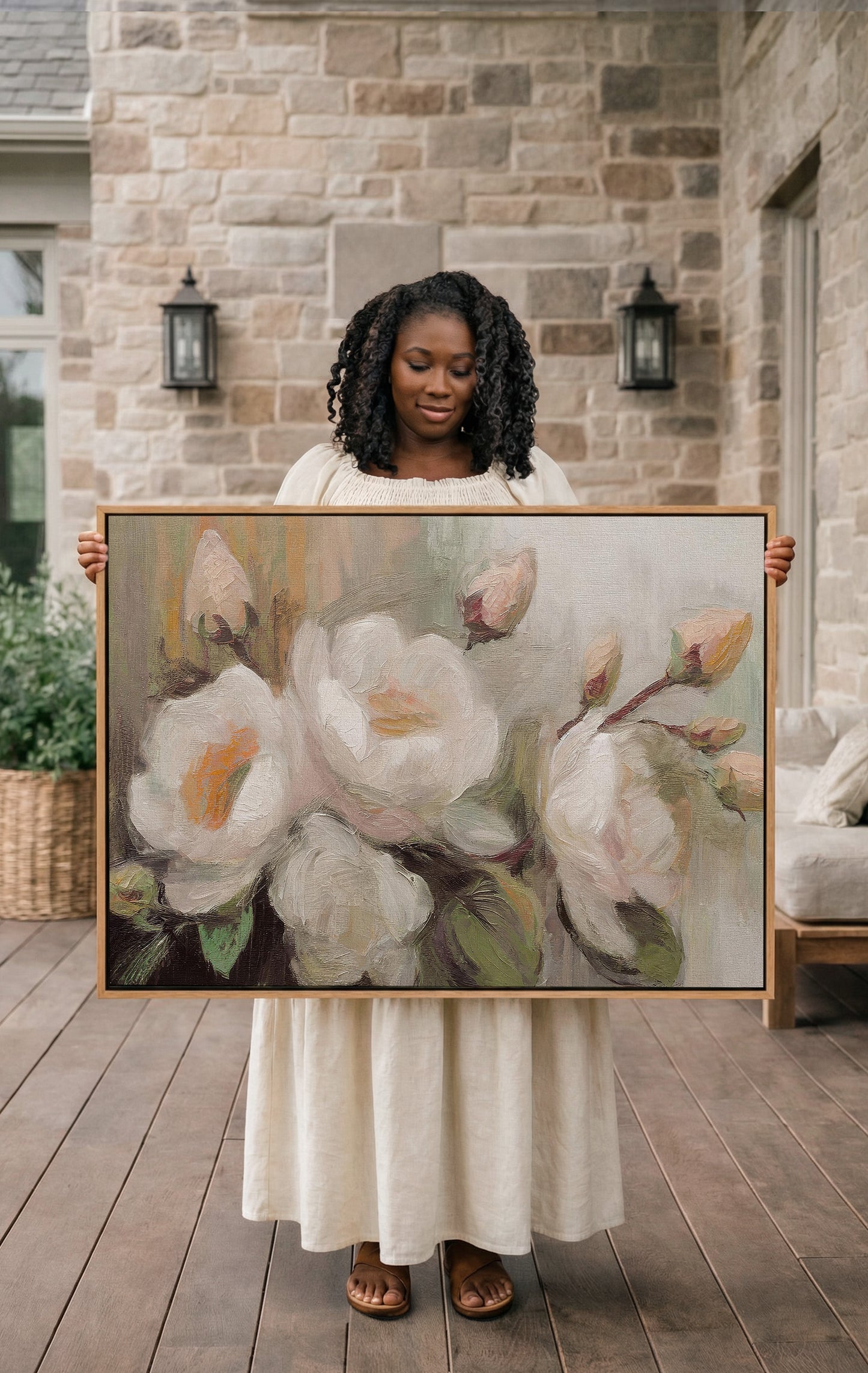 A woman in a white dress holds a large painting of white flowers on a wooden deck in front of a stone wall.