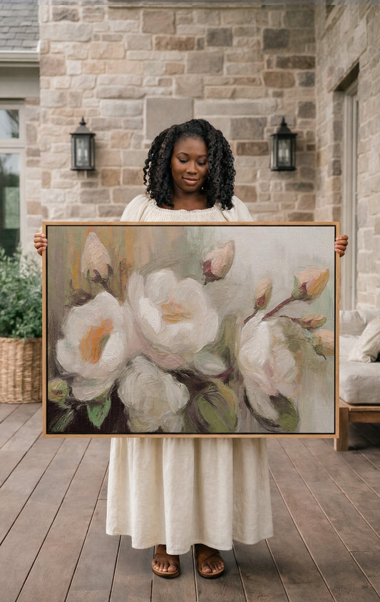A woman in a white dress holds a large painting of white flowers on a wooden deck in front of a stone wall.