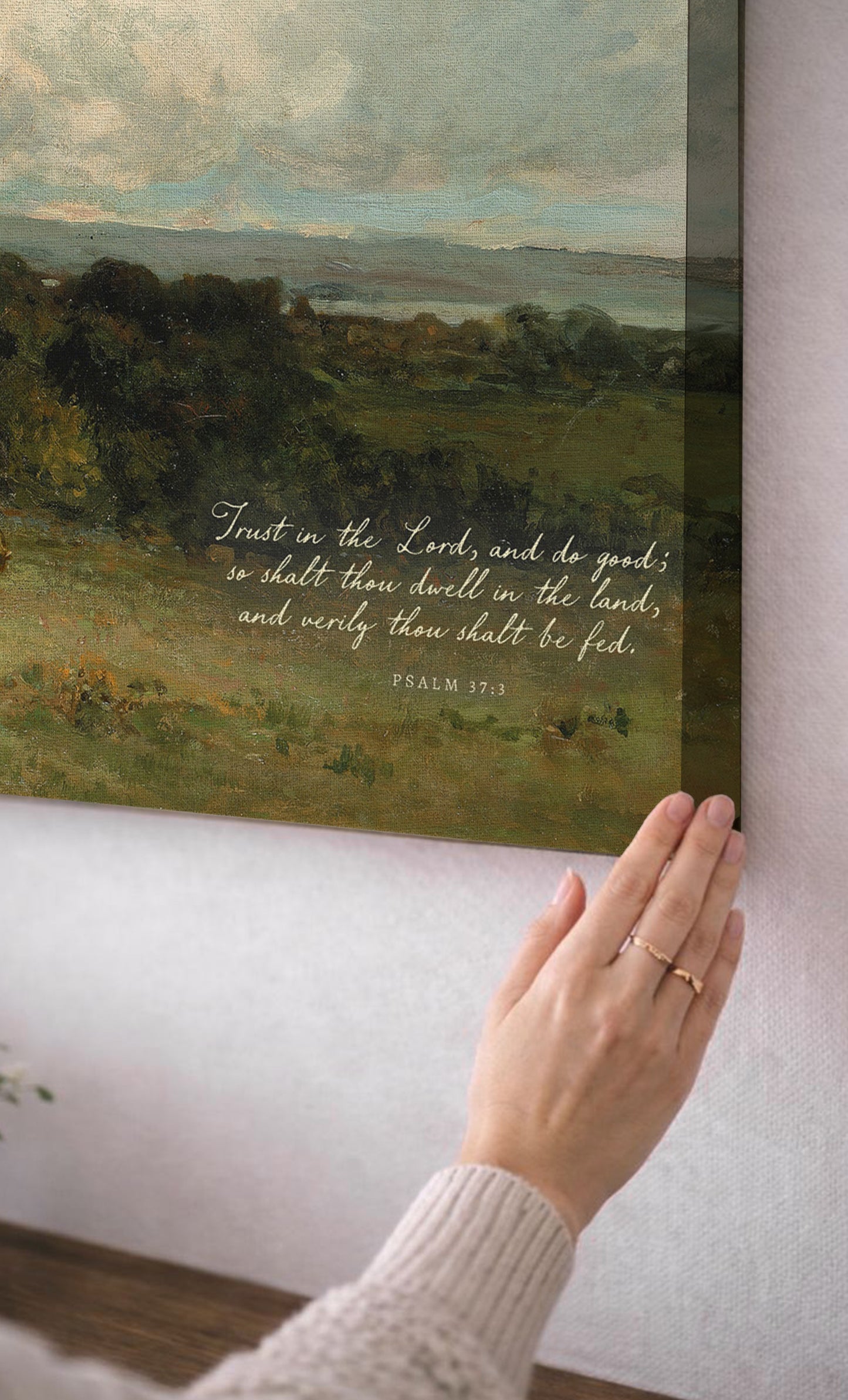A person's hand is reaching towards a framed painting of a pastoral landscape, with a Bible verse visible on the painting.