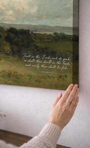 A person's hand is reaching towards a framed painting of a pastoral landscape, with a Bible verse visible on the painting.