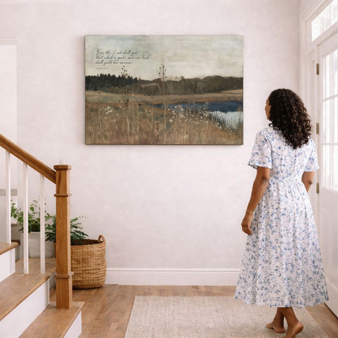 A woman in a blue and white floral dress stands in a hallway, gazing at a painting of a serene landscape hanging on the wall.