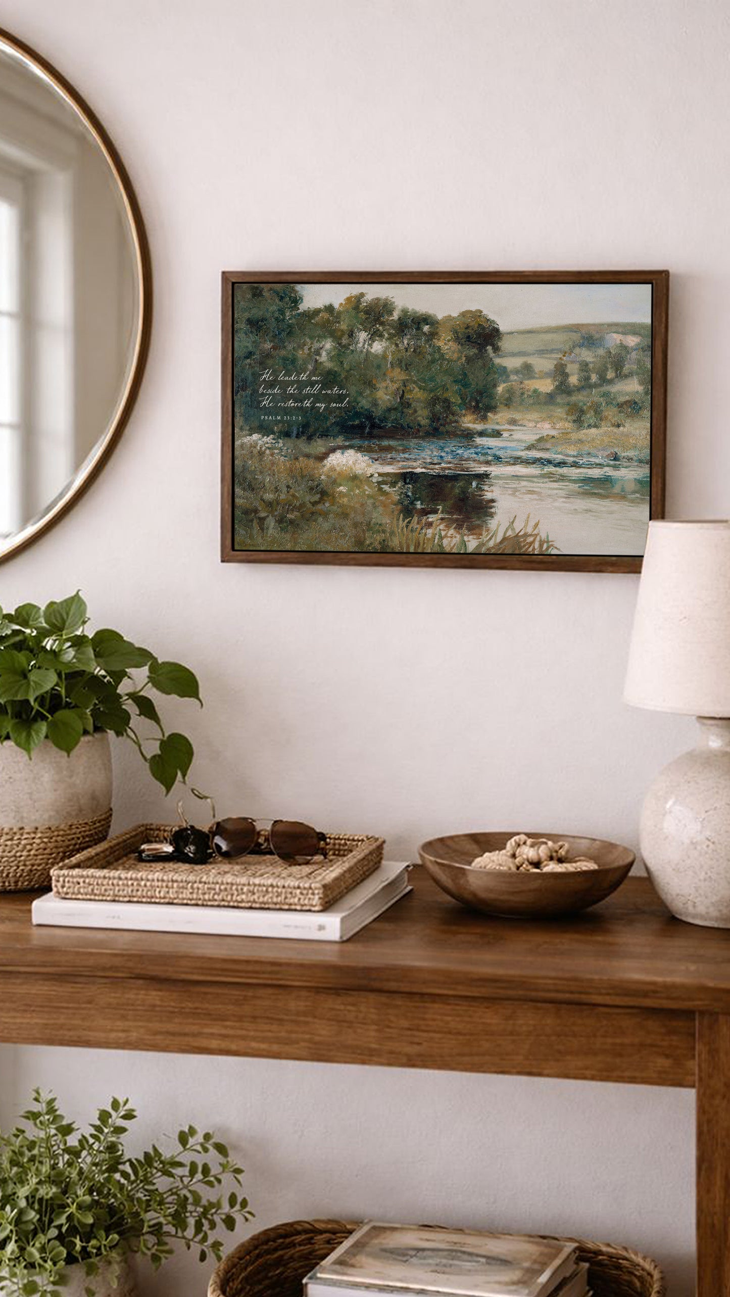 a wooden console table with various items on it, including a framed landscape painting, a lamp, a bowl, and a plant. The table is set against a white wall, and there is a round mirror above it.