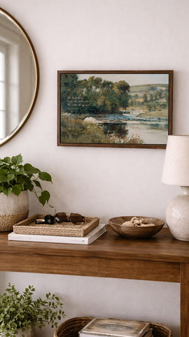 a wooden console table with various items on it, including a framed landscape painting, a lamp, a bowl, and a plant. The table is set against a white wall, and there is a round mirror above it.