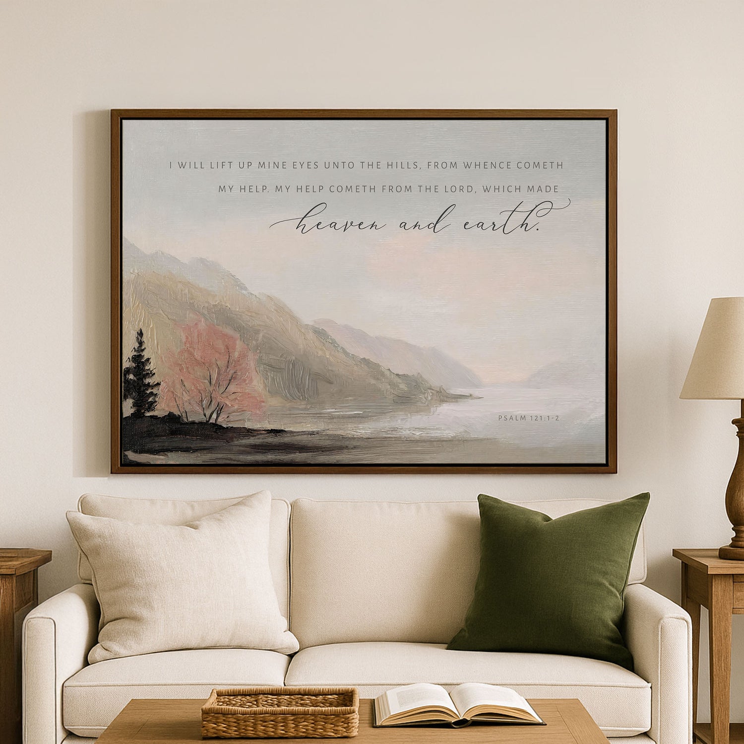 a large framed painting of a serene landscape with mountains, trees, and a body of water, hanging on a wall above a white couch with green pillows.