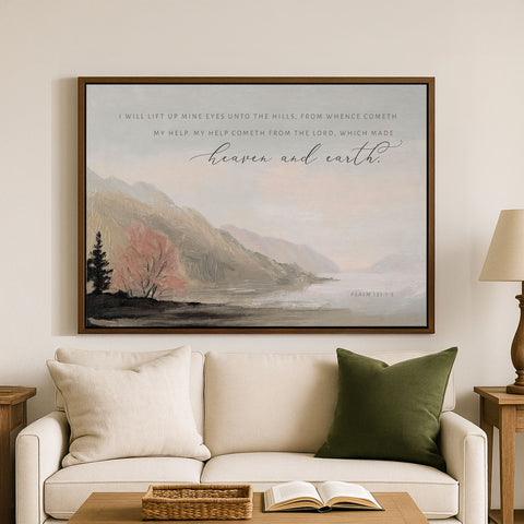 a large framed painting of a serene landscape with mountains, trees, and a body of water, hanging on a wall above a white couch with green pillows.