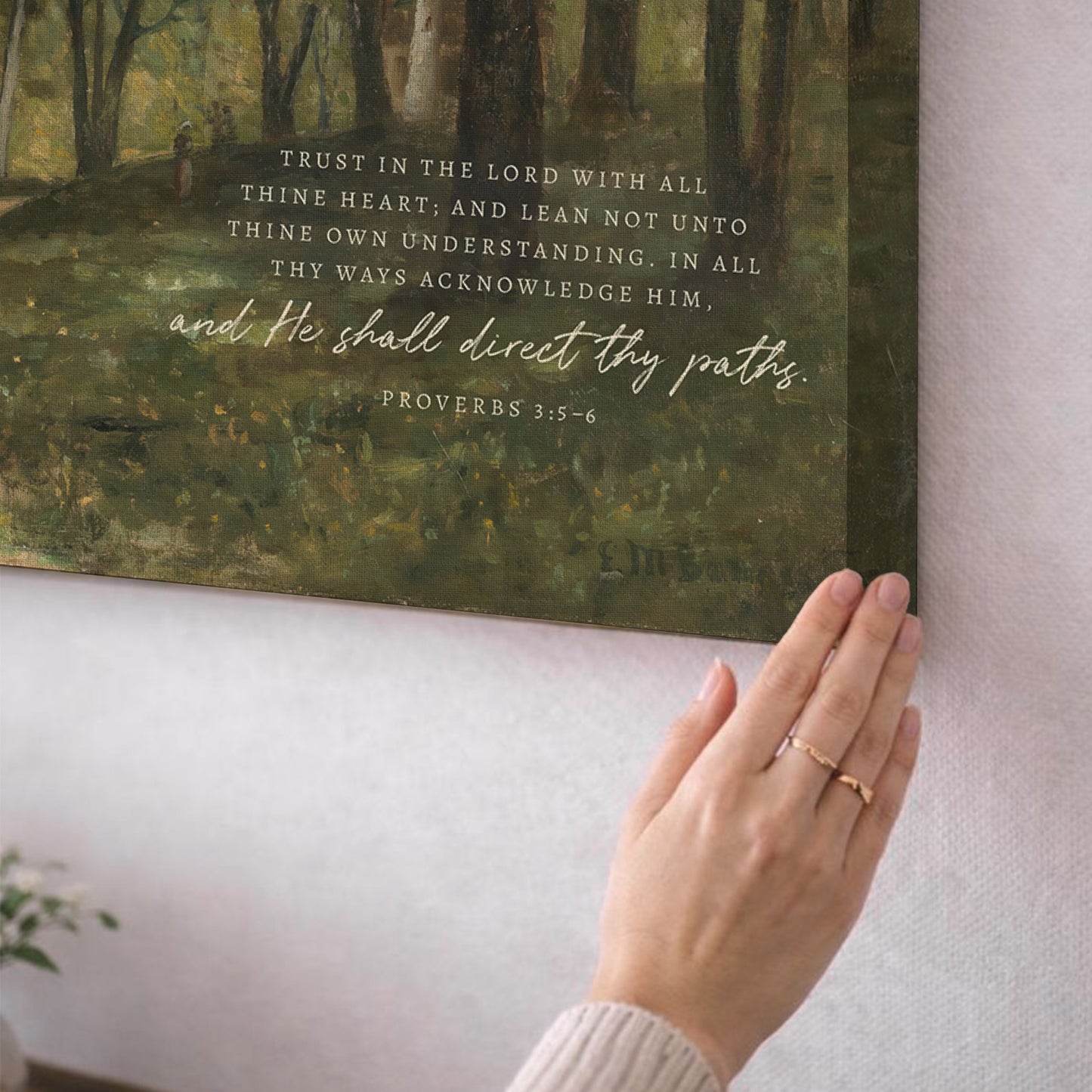 A person's hand is holding a framed Bible verse, with the text "Trust in the Lord with all thine heart, and lean not unto thine own understanding, in all thy ways acknowledging Him." The person is standing in front of a painting depicting a forest scene.