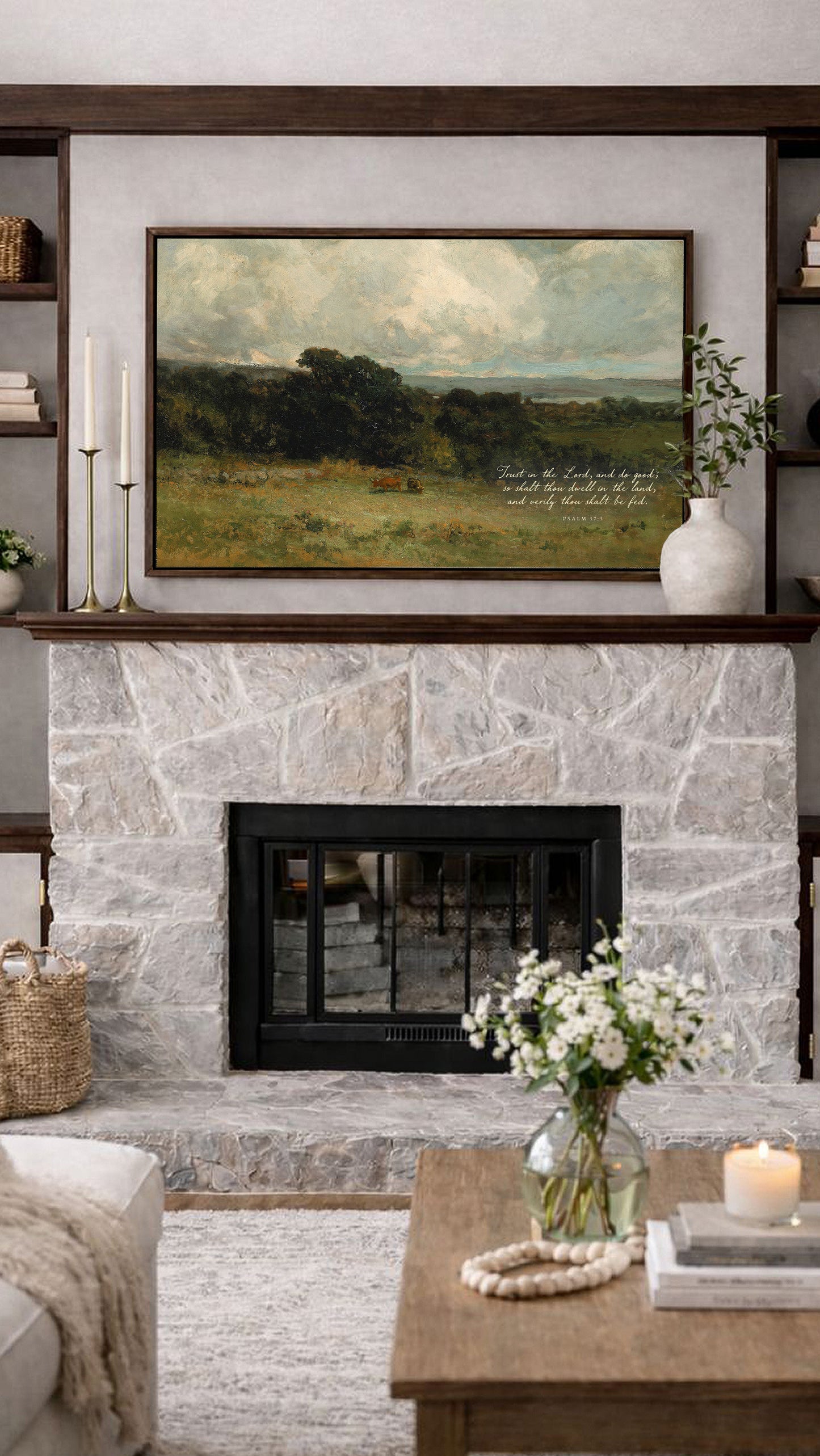 a cozy living room with a stone fireplace, a framed landscape painting above it, and a wooden coffee table with a vase of white flowers.