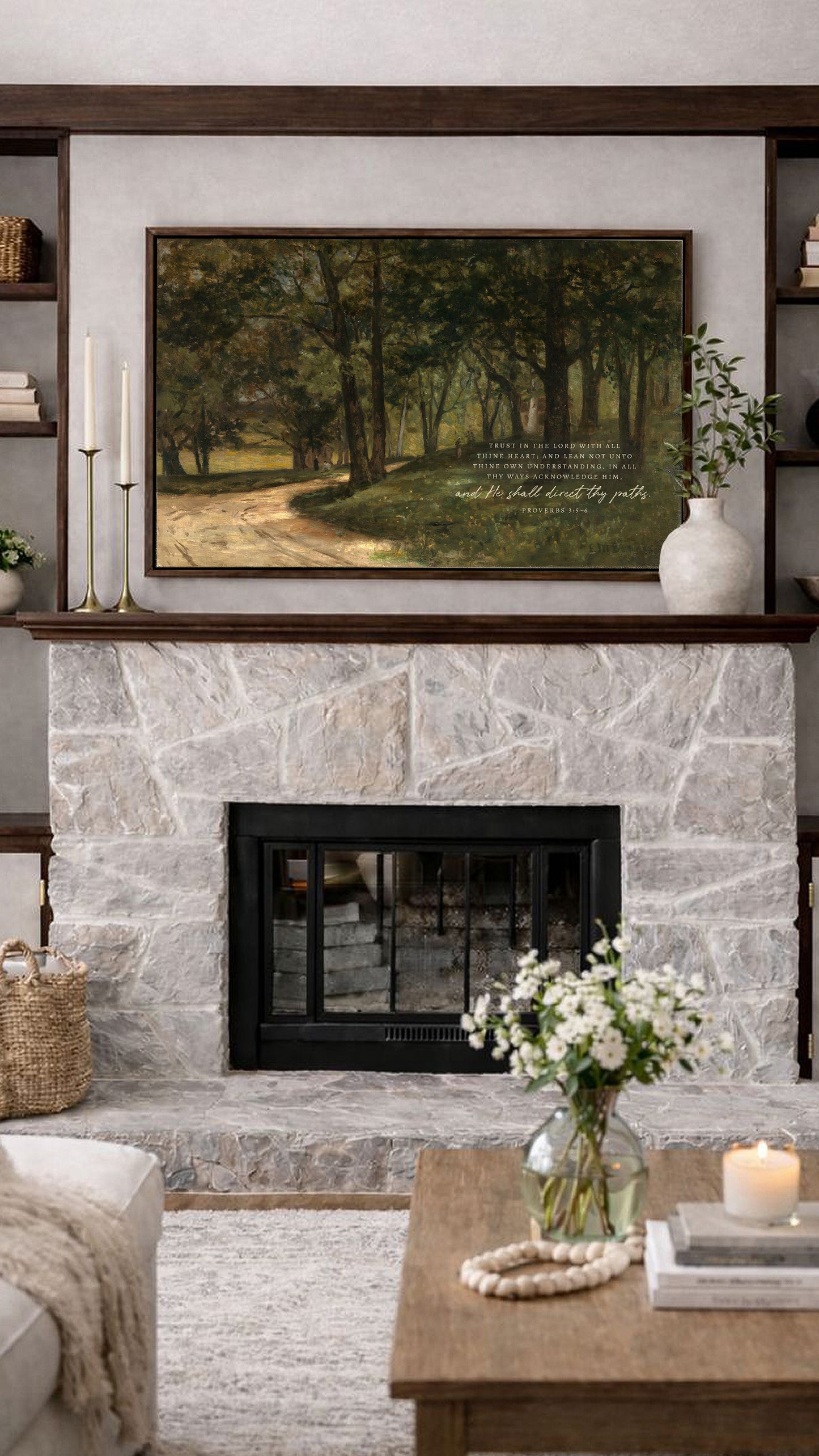 a cozy living room with a stone fireplace, a wooden coffee table, and a framed landscape painting above the fireplace. The painting depicts a serene forest scene with a winding path and lush greenery. The room is decorated with a vase of white flowers, a candle, and a bookshelf filled with various items.