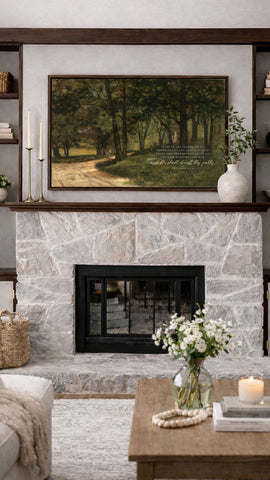 a cozy living room with a stone fireplace, a wooden coffee table, and a framed landscape painting above the fireplace. The painting depicts a serene forest scene with a winding path and lush greenery. The room is decorated with a vase of white flowers, a candle, and a bookshelf filled with various items.