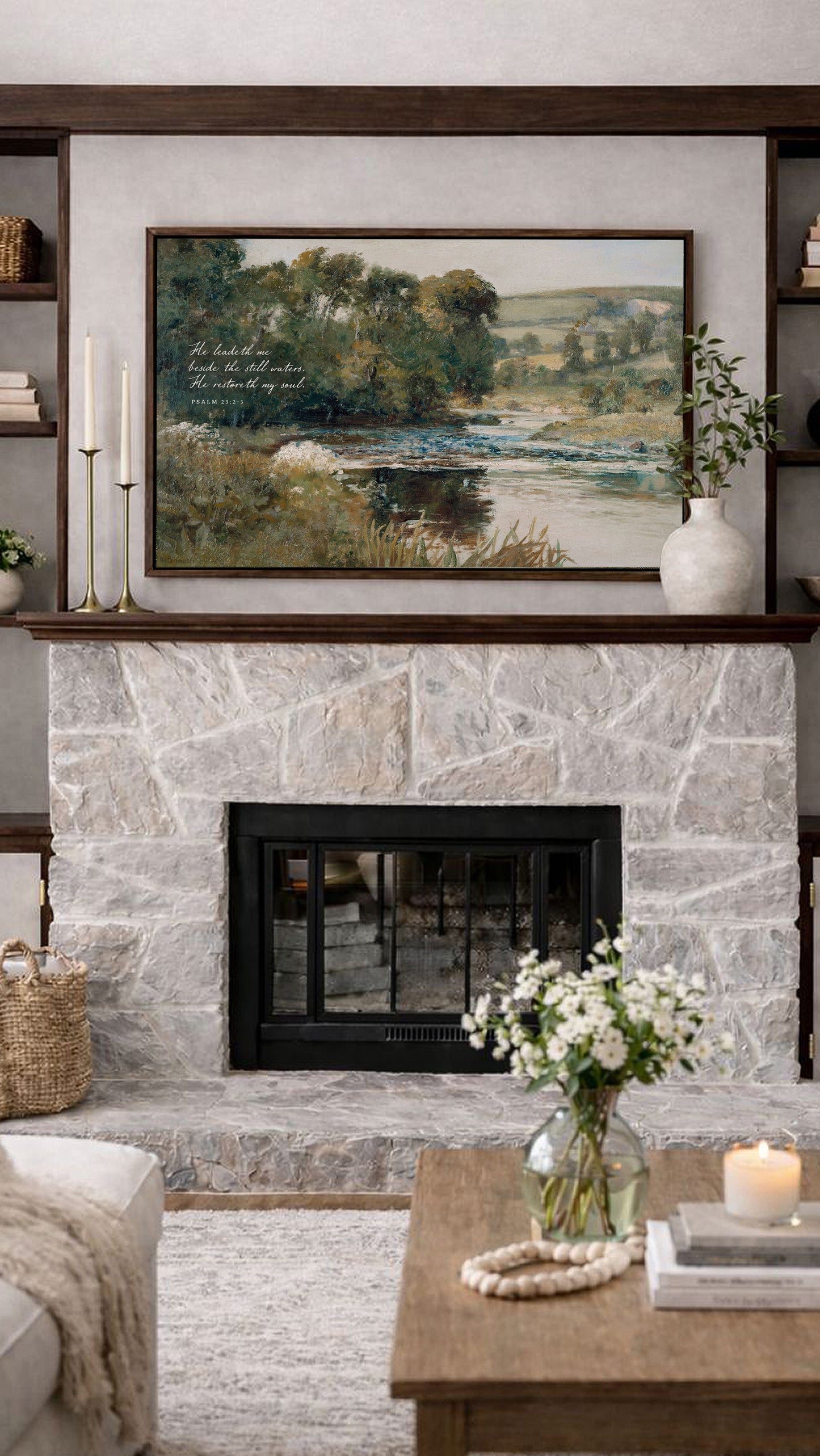 a cozy living room with a stone fireplace, a framed landscape painting above it, and a wooden coffee table with a vase of white flowers.