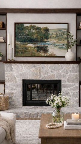 a cozy living room with a stone fireplace, a framed landscape painting above it, and a wooden coffee table with a vase of white flowers.