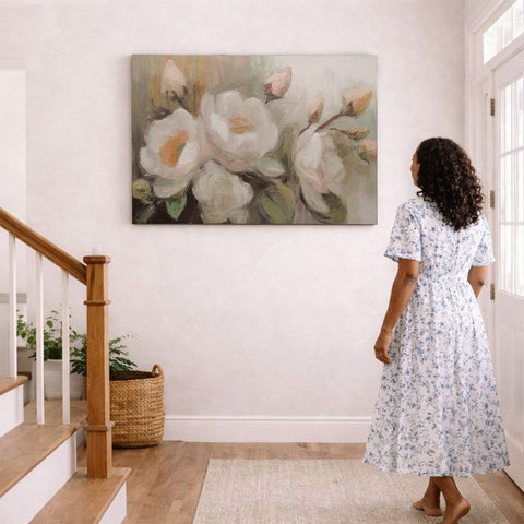 A woman in a white and blue floral dress stands in a hallway, gazing at a large painting of white and pink flowers hanging on the wall.