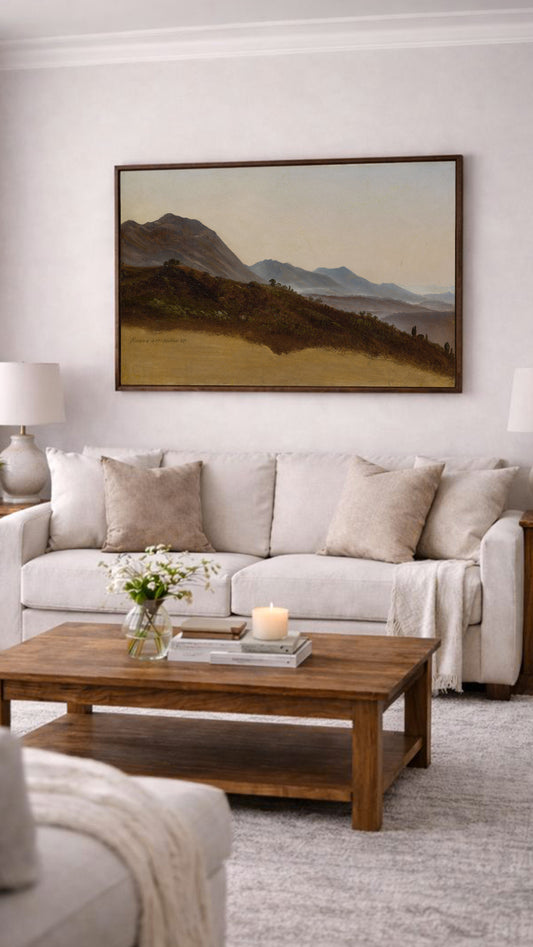 a cozy living room with a large painting of a mountain landscape hanging on the wall above a beige sofa. The sofa is adorned with two throw pillows, and a wooden coffee table sits in front of it. A vase of flowers and a lit candle on the coffee table add a touch of warmth and elegance to the space.