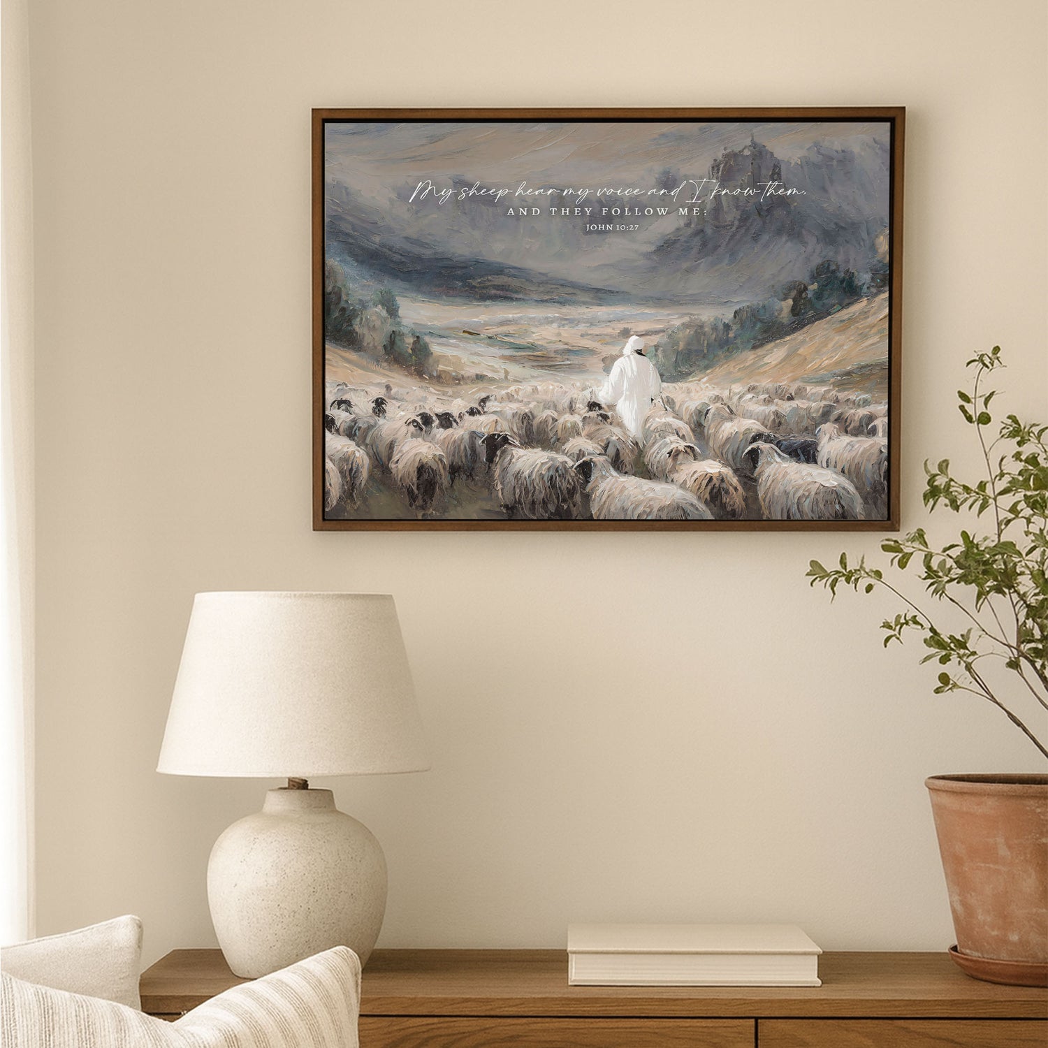 a painting of a shepherd tending to his flock, with a mountain in the background. The painting is hung on a wall above a wooden table with a lamp and a plant.