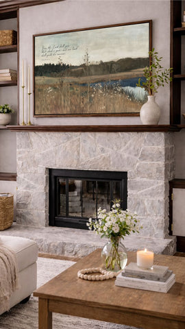 a cozy living room with a fireplace, a wooden coffee table, and a framed landscape painting above the fireplace. The painting depicts a serene countryside scene with a lake and trees. The room is decorated with a vase of white flowers, a candle, and some books, creating a warm and inviting atmosphere.
