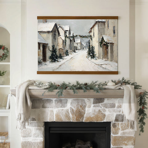a cozy fireplace adorned with a wreath and greenery, with a framed painting of a snowy village hanging above it.