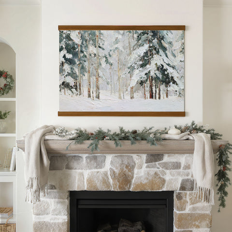 a cozy fireplace adorned with a wreath and a blanket, with a framed painting of a snowy forest hanging above it.