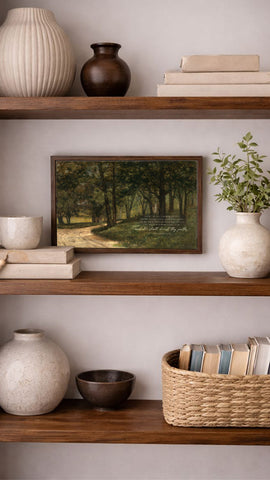 a wooden bookshelf with various decorative items and books arranged on the shelves. The bookshelf has three shelves, and the top shelf has a framed picture of a forest scene. The middle shelf has a stack of books, and the bottom shelf has a woven basket filled with books. The bookshelf also includes a white vase, a brown bowl, and a white ceramic vase.